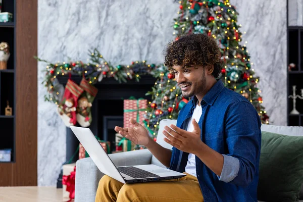 Young Christmas man sitting at home on sofa with laptop near tree decorated with Christmas decorations, Hispanic man using video call to greet friends and family remotely online.