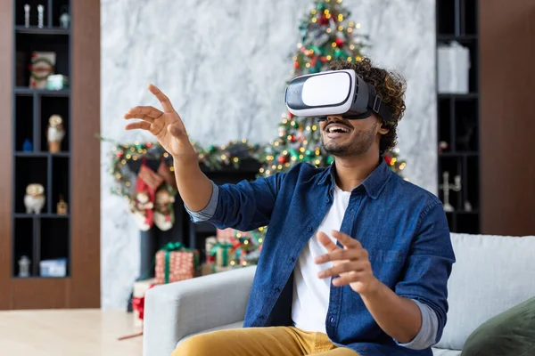 A man is using a virtual reality simulator for Christmas, sitting on a sofa in the living room of the house, near a decorated Christmas tree, on New Years holidays.