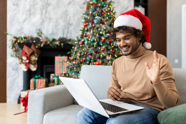 Hispanic christmas man using laptop for online video call, waving at camera greeting family and friends remotely, man in red christmas santa hat sitting on sofa in living room near christmas tree.