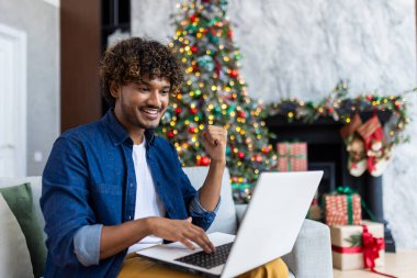 Yılbaşı ve yeni yıl tatili için oturma odasındaki koltukta uzaktan kumandayla çalışan mutlu ve başarılı bir adam. Hispanik adam elini kaldırıyor zafer jesti, ağacın yanında kazanan, başarı..