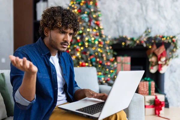 Upset man looking at laptop screen sitting at home in living room on Christmas, hispanic man frustrated writing online notification email to mail, near Christmas tree on New Year holidays.