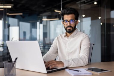 Ofiste çalışan Hintli bir adamın portresi, bir dizüstü bilgisayarın arkasında oturuyor ve özgüvenle kameraya bakıyor..
