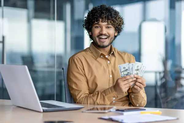 Portrait of a happy Indian man sitting in the office at a desk with a laptop and holding cash money in his hands, smiling and happy at the camera.