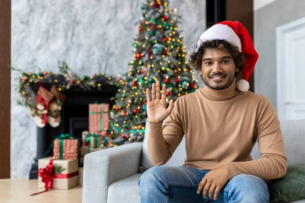 Web camera view, online video call for the new year. A man in a red Santa hat smiles and looks at the camera, waves hand to say hello, sits on a sofa near a Christmas tree, records a greeting video.