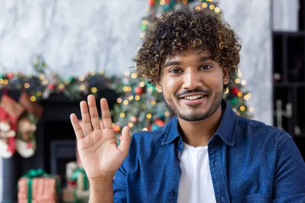 Close-up man waving at web camera, smiling and happy greeting, hispanic man on christmas recording online greeting video for family and friends, sitting in living room of house on sofa near tree.