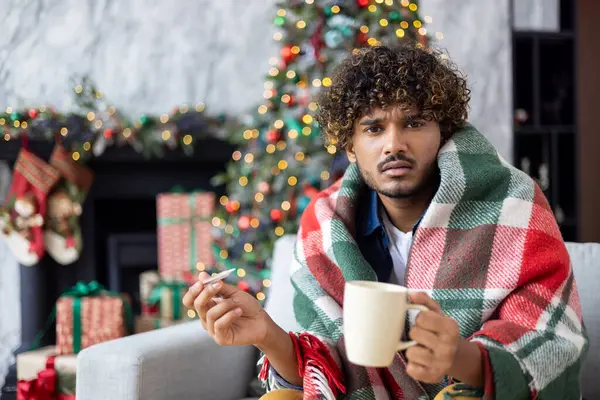 Portrait of sick man at home on sofa, man celebrating new year and christmas, sitting on winter day in living room close up and looking at camera, near decorated Christmas tree, flu and cold.