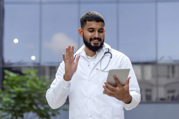 A smiling young male Indian doctor stands on the street near the hospital in a white coat and communicates via video call with patients, consults remotely online, greets and waves at the camera.