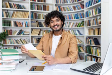 Üniversite kütüphanesinde bir masada oturan genç Müslüman bir erkek öğrencinin portresi ve kameraya gülümseyen ve işaret eden bir mektup açılıyor..