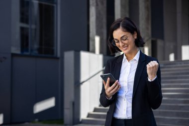 İş elbiseli genç ve güzel bir iş kadını elinde telefonla şehirde yürüyor. Bir kadın, zafer kazandığını bildiren bir başvuru formu kullanıyor. Zaferi ve başarıyı kutluyor..