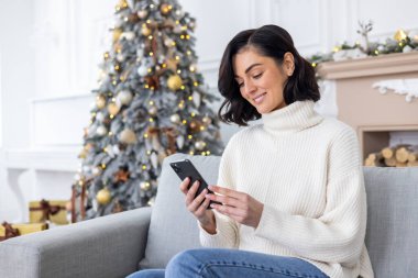Genç ve güzel bir kadın oturma odasındaki koltukta süslü Noel ağacının yanında oturuyor, gülümsüyor ve telefon ediyor, akıllı telefonda mesaj yazıyor, yeni yılı ve Noel 'i kutluyor..