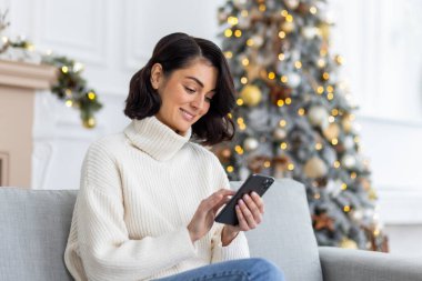 Genç ve güzel bir kadın oturma odasındaki koltukta süslü Noel ağacının yanında oturuyor, gülümsüyor ve telefon ediyor, akıllı telefonda mesaj yazıyor, yeni yılı ve Noel 'i kutluyor..
