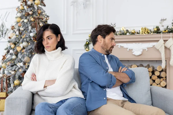 Frustrated and upset family couple sitting on sofa in living room on christmas, man and woman quarreling not talking, celebrating new year and christmas near tree on winter day.