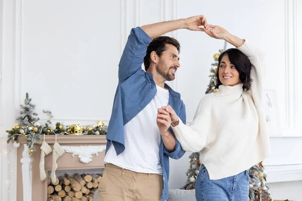 Young married couple man and woman celebrating new year and christmas together, dancing in the living room of the house near the Christmas tree and hugging smiling.