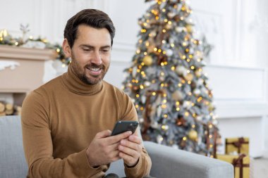 Yılbaşı tatillerinde ve hafta sonlarında Noel ağacının yanındaki kanepede oturan gülümseyen genç bir adam cep telefonu kullanır. Yakın plan fotoğraf..