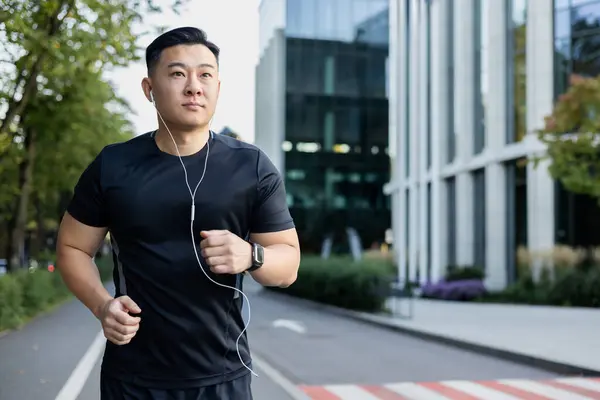 Young Asian male athlete runs in the city on the street in headphones, listens to music, rests.