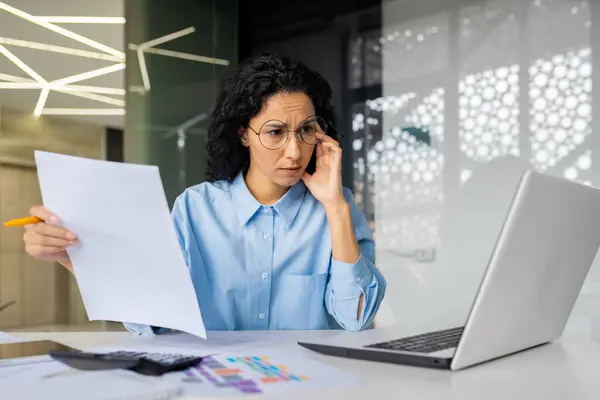 Upset and disappointed businesswoman behind paperwork, financier checking papers, contracts, documents and reports, female worker inside office working with laptop, unhappy with achievement results.