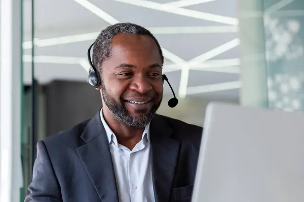 Mature experienced man in business suit with headset phone, working inside office with laptop, businessman online customer support worker, consulting customers remotely, selling in telestore.