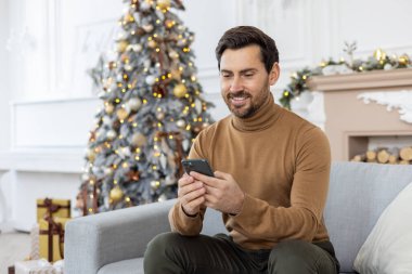 Genç bir adam kış tatilinde Noel ağacının yanındaki koltukta oturuyor ve cep telefonu kullanıyor..