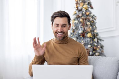 Yeni yıl tatili ile ilgili bir video görüşmesinde genç bir adamın konuşmasının ve selamlaşmasının yakın plan fotoğrafı. Noel ağacının yanında dizüstü bilgisayarla kanepede oturmak..