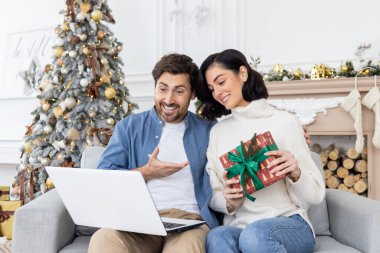 Genç bir çift, bir erkek ve bir kadın, evde kanepede birlikte oturuyorlar, bir Noel hediyesi tutuyorlar ve dizüstü bilgisayardan telefonla konuşuyorlar. İnternette mutlu tatiller.