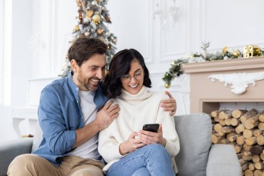 Genç ve mutlu bir çift, evlerinde oturmuş Noel ağacının yanında sarılıyor, cep telefonu kullanıyor, internetten alışveriş yapıyor, video izliyor..