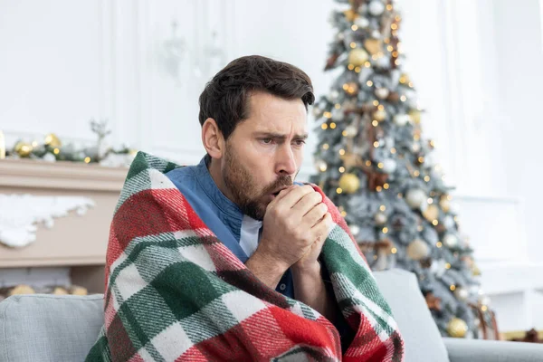 Close-up photo of a young man sitting at home on the sofa covered with a blanket, blowing warm air from his mouth into his hands, freezing from the cold.