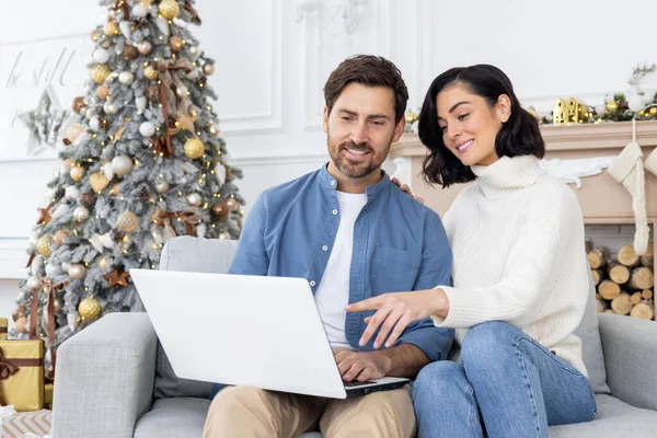 Portrait of a young family, a married couple, a man and a woman sitting at home on New Years holidays doing online shopping. Sitting on the sofa, holding a laptop, chatting and choosing a product.