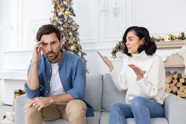 Conflict of a young couple, family on New Years holidays. A young man and a woman are sitting on the couch and arguing. The wife screams and accuses the husband, the husband holds his head.