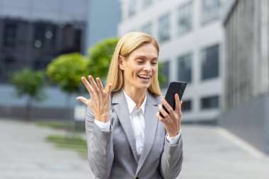 Başarılı, olgun, neşeli iş kadını internette iyi sonuçlar alan bir mesaj aldı. Telefonla iyi sonuçlar elde etti. Kadın patron uygulamayı akıllı telefonda kullanıyor, ofis binasının önünde takım elbiseyle yürüyor..