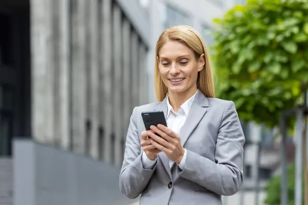 stock image Successful woman boss walks outside office building outdoors, in business suit, businesswoman uses app on phone, smiles contentedly reads internet pages, browses news, writes text message, call.