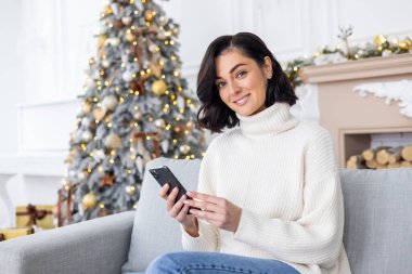 Yılbaşı arifesinde evdeki kanepede oturan genç ve güzel bir kadının portresi Noel ağacının yanında cep telefonuyla kameraya gülümsüyor..