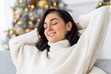 İşten sonra evde dinlenen ve Noel ve yeni yıl tatillerinde tatilde olan gülümseyen genç bir kadın. Elleri başının arkasında kanepede oturur, gözlerini kapatır. Yakın plan fotoğraf..