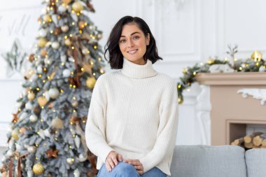 Gülümseyen genç bir kadının portresi. Beyaz bir kazakla Noel ağacının yanında oturuyor ve kendine güvenerek kameraya bakıyor..