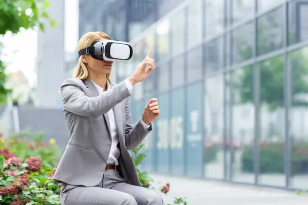 Businesswoman outside office building uses virtual reality glasses, female boss sits on a bench in the daytime in a business suit.