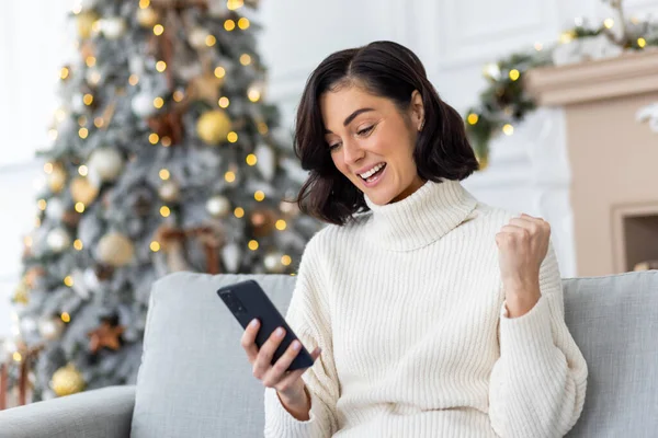 Happy young woman sitting at home on sofa near Christmas tree and using mobile phone, happy with success and good news for New Year holidays, showing victory gesture with hand.