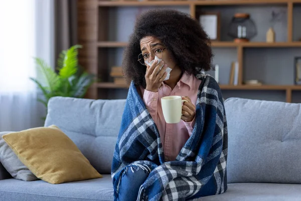 Sick cold woman sitting on sofa in living room, African American woman under blanket, has runny nose and high body temperature, sneezes and drinks hot drink with medicine.
