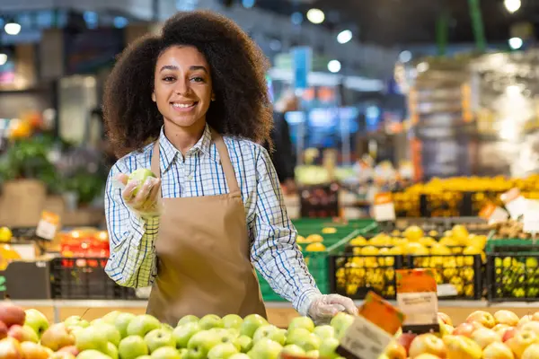 Portrait of a young saleswoman in a supermarket, grocery store, a smiling saleswoman looking at the camera with satisfaction, looking at apples, a Hispanic woman with curly hair in an apron.