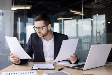 Genç bir ofis çalışanı masadaki ofiste oturuyor, elinde kağıtlarla, şaşırtıcı bir şekilde belgelere ve faturalara bakıyor..