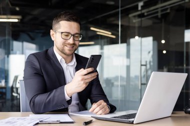 Gülümseyen genç bir adam ofiste dizüstü bilgisayarla çalışıyor ve cep telefonu kullanıyor..