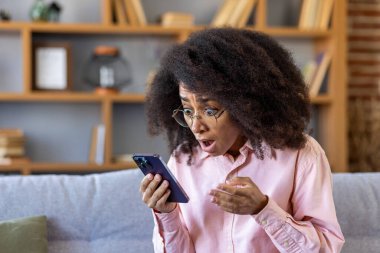 Hüzünlü kadın telefonda flaş haber aldı, Afrikalı Amerikalı kadın internette kötü haber okuyor, akıllı telefondaki uygulamayı kullanıyor, evdeki oturma odasında kanepede oturuyor..