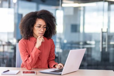 İş yerindeki başarılı ciddi kadın, kırmızı gömlekli iş kadını dizüstü bilgisayarla çalışıyor, projenin finansal çözümünü düşünüyor.