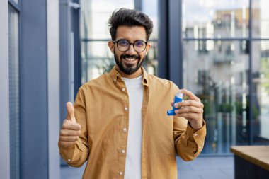 Neşeli ve gülümseyen adam iyileşti, kameraya bakıyor nefes alıp verişini kolaylaştırmak için astım spreyini gösteriyor, dışarıda ofis binasında yürüyor..