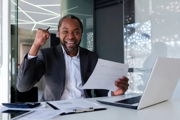 Happy successful banker smiling and looking at camera financier investor behind paper work, man celebrating victory success and triumph, businessman inside office at work with laptop, holding report.