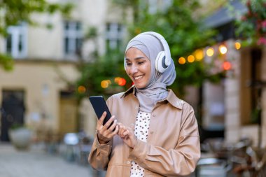 Şehirde yürüyen genç, güzel bir kadın, akşam tesettürlü Müslüman bir kadın kulaklıklarla yürür akıllı telefon uygulaması kullanarak neşeyle gülerek müzik dinler..