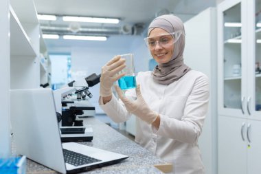 Laboratuvarda tesettürlü ve beyaz önlüklü olgun bir Müslüman kadın çalışıyor, kadın tatmin olmuş bir şekilde gülümsüyor, aletlerle deney yapıyor, dizüstü bilgisayar ve mikroskop kullanıyor..