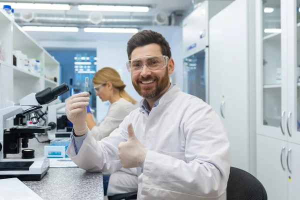 Satisfied with research results, scientist lab assistant shows thumbs up to camera, mature experienced lab worker uses microscope inside building