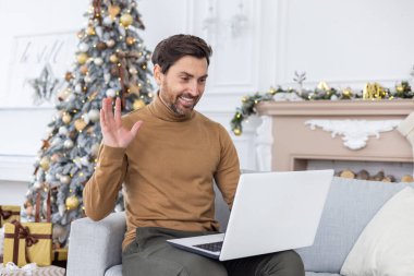 Genç bir adam, yeni yıl ve Noel tatillerinde dizüstü bilgisayarında video görüşmesinde konuşuyor. Evde kanepede oturup kameraya el sallamak ve gülümsemek..