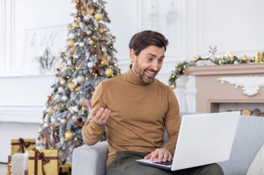 Genç bir adam evdeki kanepede otururken dizüstü bilgisayardaki bir video görüşmesinde konuşuyor. Gülümsüyor ve kameraya işaret ediyor..