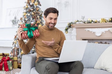 Gülümseyen genç bir adam dizüstü bilgisayardaki bir video görüşmesinde konuşuyor. Kanepede oturup kameraya yılbaşı hediyesi göstermek, Noel tatili için tebrikler..