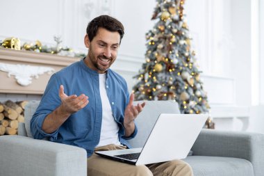 Genç adam dizüstü bilgisayarla konuşuyor evdeki kanepede oturuyor, gülümsüyor ve elleriyle el kol hareketi yapıyor..